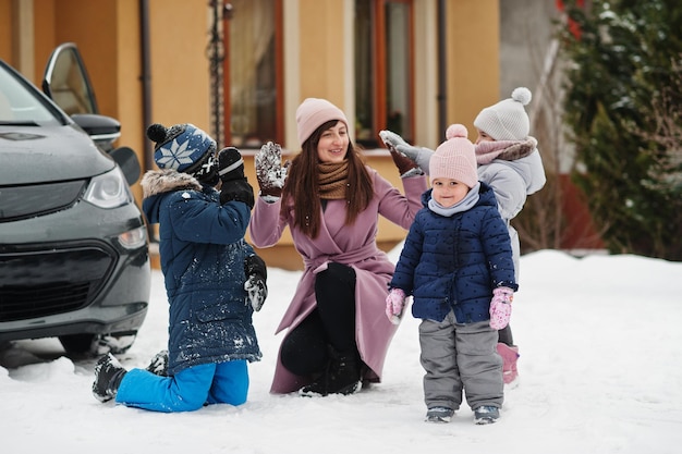 Young mother play with kids against electric car in the yard of her house at winter