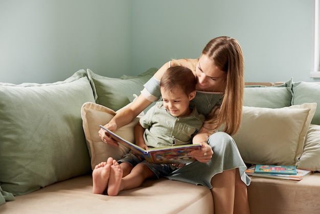 Young mother, read a book to her child, boy in the living room of their home, rays of sun going through the window