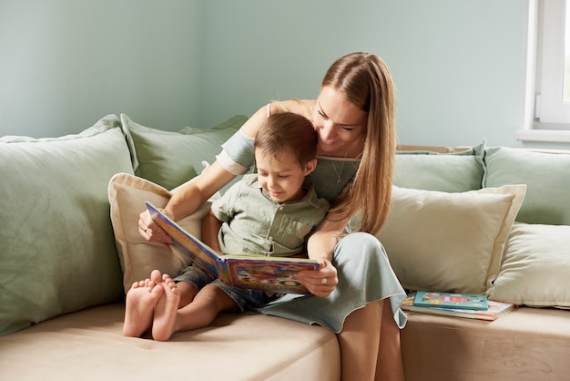 Young mother, read a book to her child, boy in the living room of their home, rays of sun going through the window