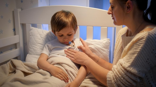 Young mother sitting next to her sick son and measuring him body temperature