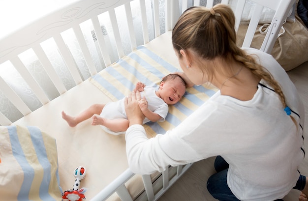 Young mother sitting near the cradle and holding baby's hand