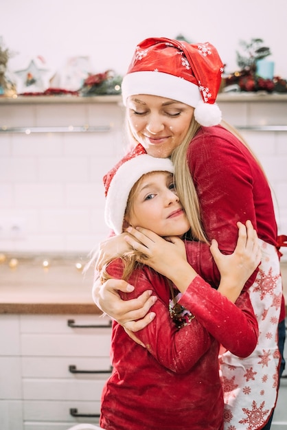 A young mother stands with her teenage child in red sweaters in the kitchen hugging in santa hats