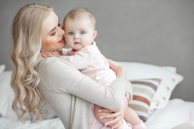 Young mother taking care of her little baby girl. Beautiful mom and her daughter indoors in the bedroom. Loving family. Attractive mum holding her child.