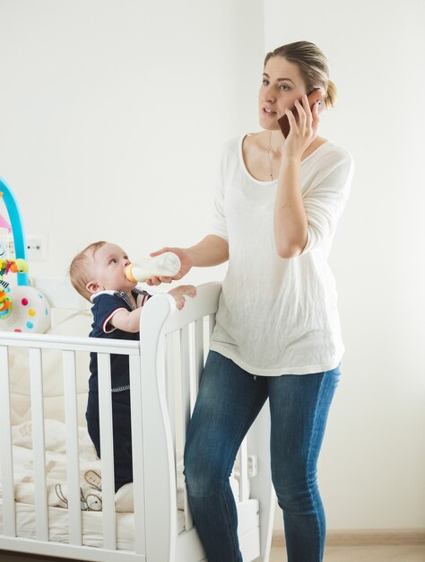 Young mother talking by phone and feeding her baby from bottle