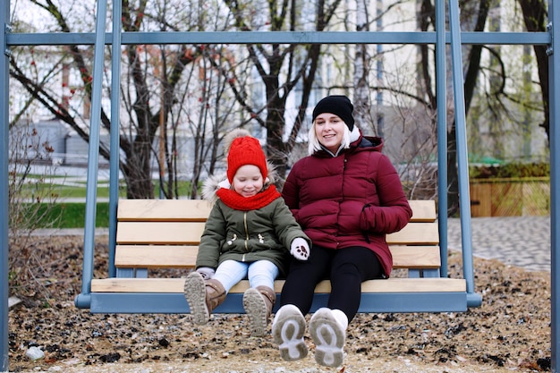 Young mother talking to her cute little daughter outdoors sitting together in warm clothes