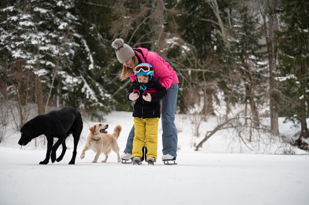 Photo young mother teaching her toddler child how to ice skate on a beautiful natural frozen lake in a snowy winter nature with their dogs around them