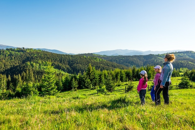 Young mother and two little daughters travelers stand on a slope with a gorgeous view of the hills covered with dense fir forest against the blue sky on sunny warm summer day. Family tourism concept