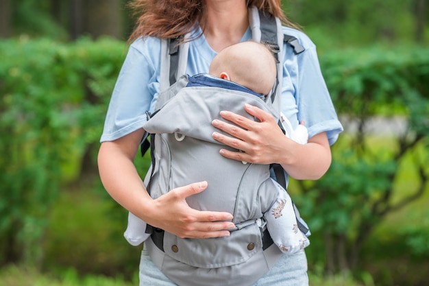 A young mother walks with a baby sleeping in a sling backpack in a green summer park