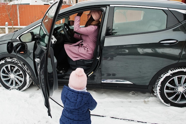 Young mother with baby girl charging electric car in the yard of her house at winter