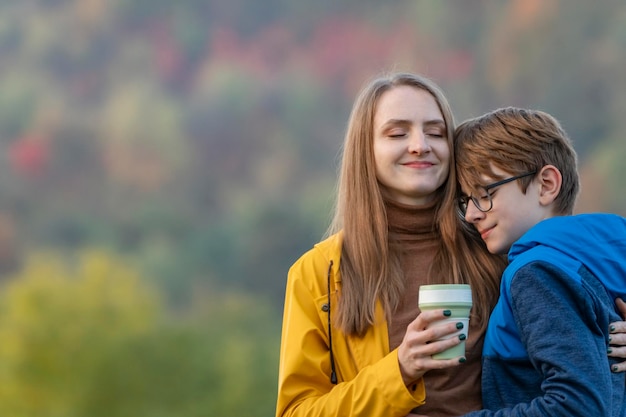 Young mother with cup of hot drink hugs son in nature Mom and child happily spend autumn day outdoors