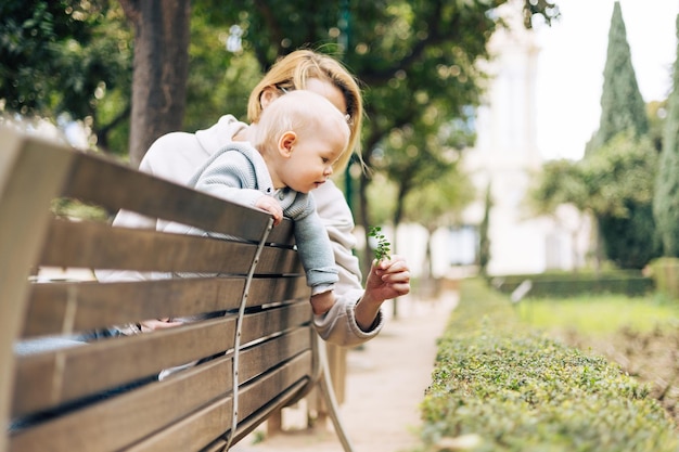 Young mother with her cute infant baby boy child leaning over back of wooden bench towards bushes in city park holding and observing green plant with young leaves and learn about life and nature