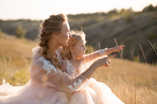 Young mother with a little daughter in pink dresses are sitting in the field