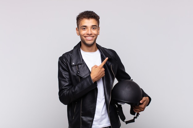 Young motorbike rider smiling cheerfully, feeling happy and pointing to the side and upwards, showing object in copy space