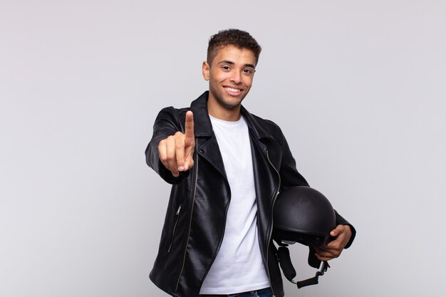 Young motorbike rider smiling proudly and confidently making number one pose triumphantly, feeling like a leader