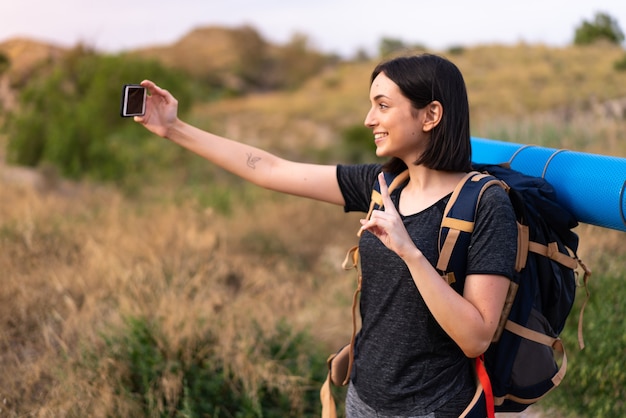 Young mountaineer girl taking a selfie with the mobile with a big backpack at outdoors