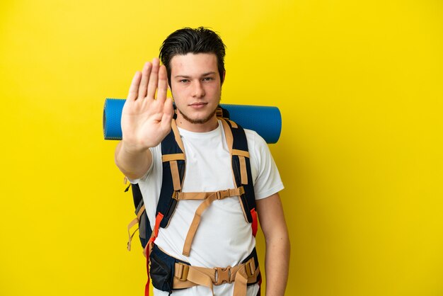 Young mountaineer Russian man with a big backpack isolated on yellow background making stop gesture