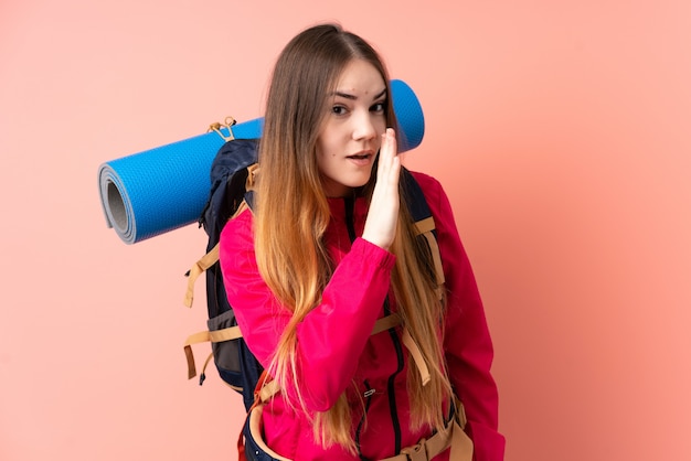 Young mountaineer woman with a big backpack isolated on pink wall whispering something