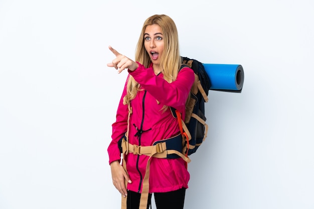 Young mountaineer woman with a big backpack over isolated white background pointing away