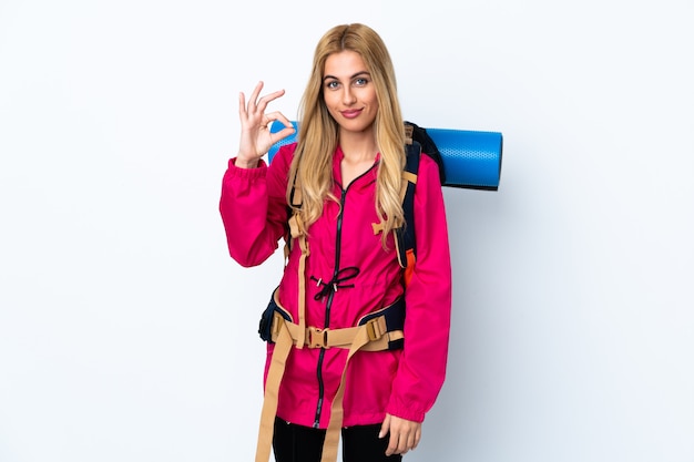 Young mountaineer woman with a big backpack over isolated white space showing an ok sign with fingers