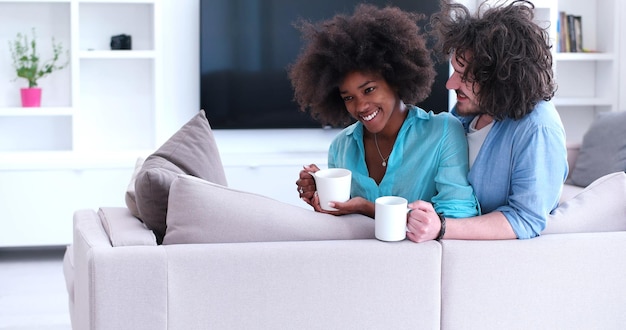 young multiethnic couple sitting on sofa at home drinking coffe, talking, smiling.