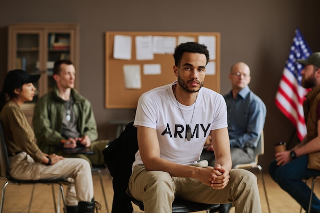 Young multiethnic unhappy man in casualwear sitting against support group