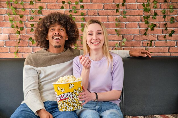 Young multiracial couple sitting on the sofa at home watching tv and smiling while eating popcorn