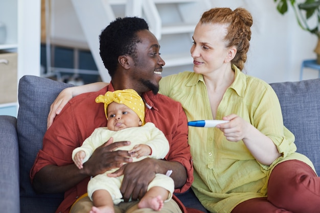 Young multiracial couple sitting on sofa with baby and making pregnancy test