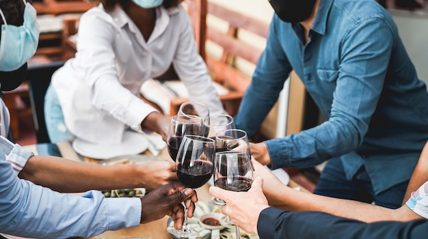 Young multiracial people cheering with wine while wearing protective masks - Social distance concept - Focus on bottom white hand