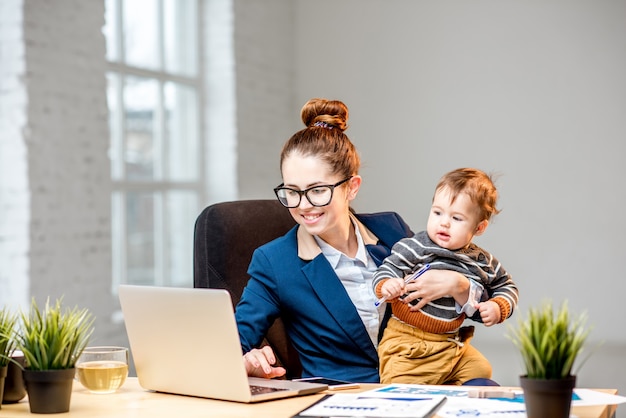 Young multitasking businessmam dressed in the suit working with laptop and documents sitting with her baby son at the office