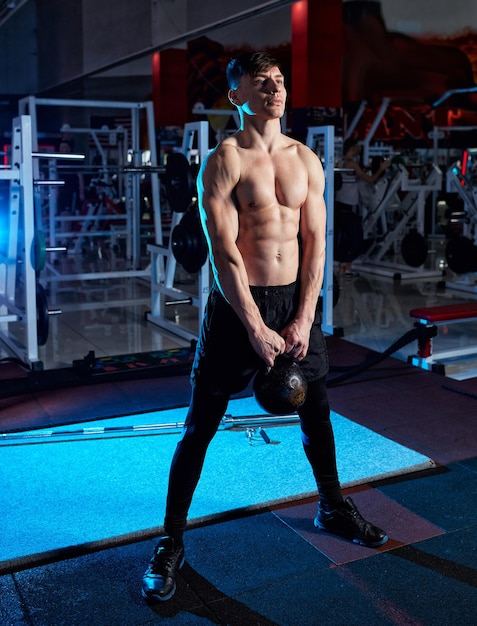 Young muscular man with naked torso working out in gym Athletic male adult exercising with kettle bell Fitness sports concept