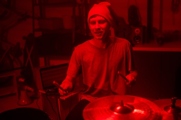 Young musician in beanie and t-shirt hitting cymbals and drums with drumsticks while sitting