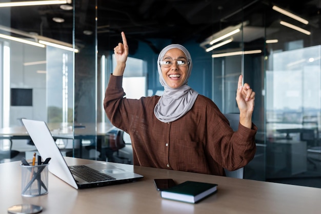 A young muslim business woman in a hijab sits in the office at the table and dances she looks