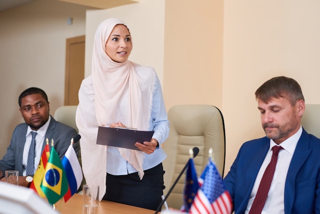 Young muslim female in hijab making report at conference while standing in front of audience in the hall