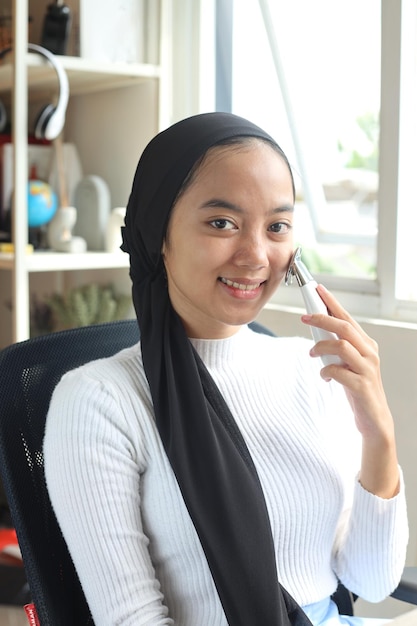 Young muslim teen girl in casual style doing facial spa using beauty tool at home.