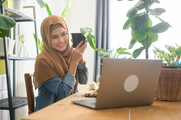 A young muslim woman entrepreneur working with laptop presents houseplants during online live stream at home