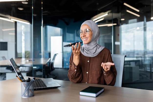 Young muslim woman freelancer in hijab sitting at office desk in front of laptop and talking on