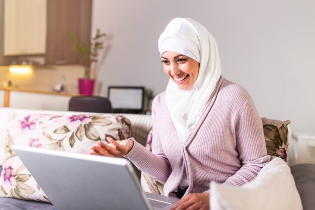 Young Muslim woman having video call via laptop at home. Happy smiling Muslim woman sitting on sofa