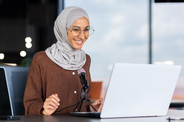 A young muslim woman in a hijab is sitting in an office in front of a laptop and a microphone