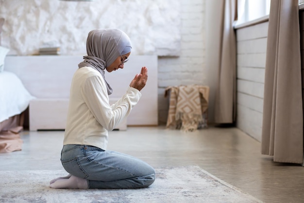 A young muslim woman in a hijab sits at home on her knees and folded her hands closed her eyes