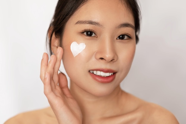 Young naked chinese lady with cream on her cheek smiling at camera using nourishing or hydrating