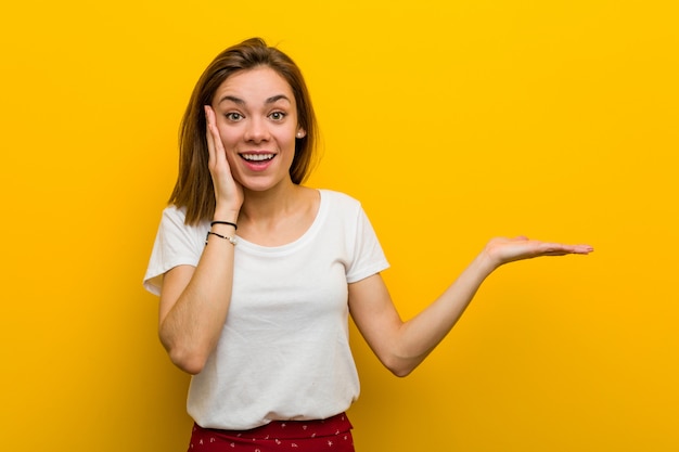 Young natural caucasian woman holds copyspace a palm, keep hand over cheek. Amazed and delighted.