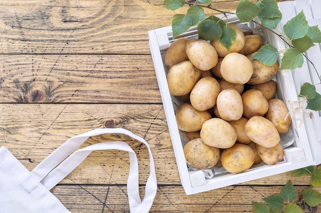 Young natural farm potatoes in a white wooden box and leaves and textile bag on a wooden background