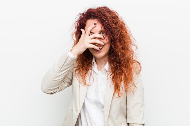 Young natural redhead business woman isolated against white blink at the camera through fingers, embarrassed covering face.