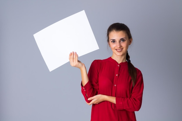 Young nice woman in red blouse with plait holding white sheet of paper on grey background in studio