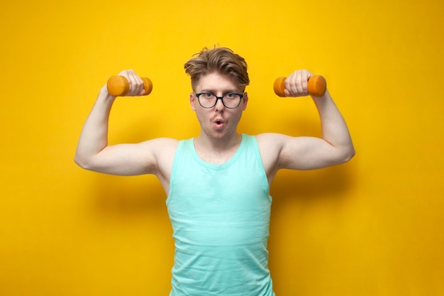 Photo young nonathletic guy with glasses raises small dumbbells on a yellow background and shows surprise