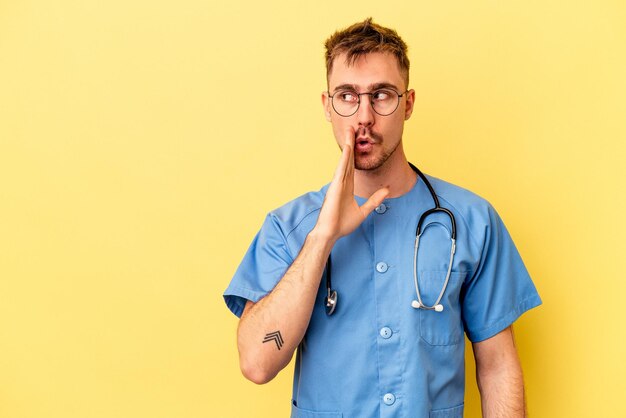 Young nurse caucasian man isolated on yellow background is saying a secret hot braking news and looking aside