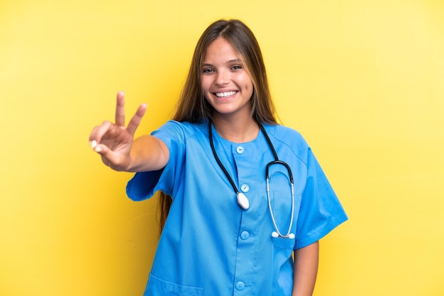 Young nurse caucasian woman isolated on yellow background smiling and showing victory sign