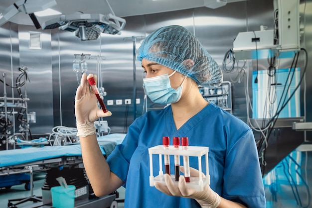 Young nurse doctor in protective uniform glove hat and mask looks at blood samples in operating room hospital