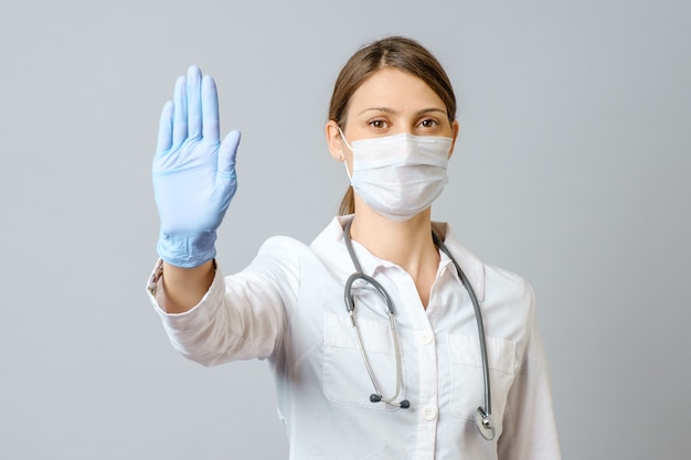 Young nurse or doctor woman wearing medical face mask and disposable gloves showing stop gesture isolated against gray wall