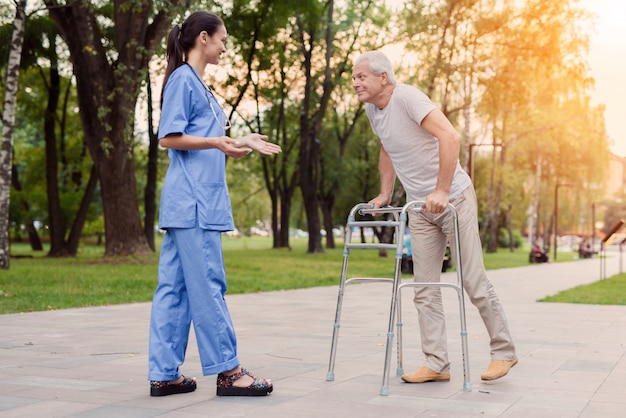 A young nurse is standing in the park and helps elderly man